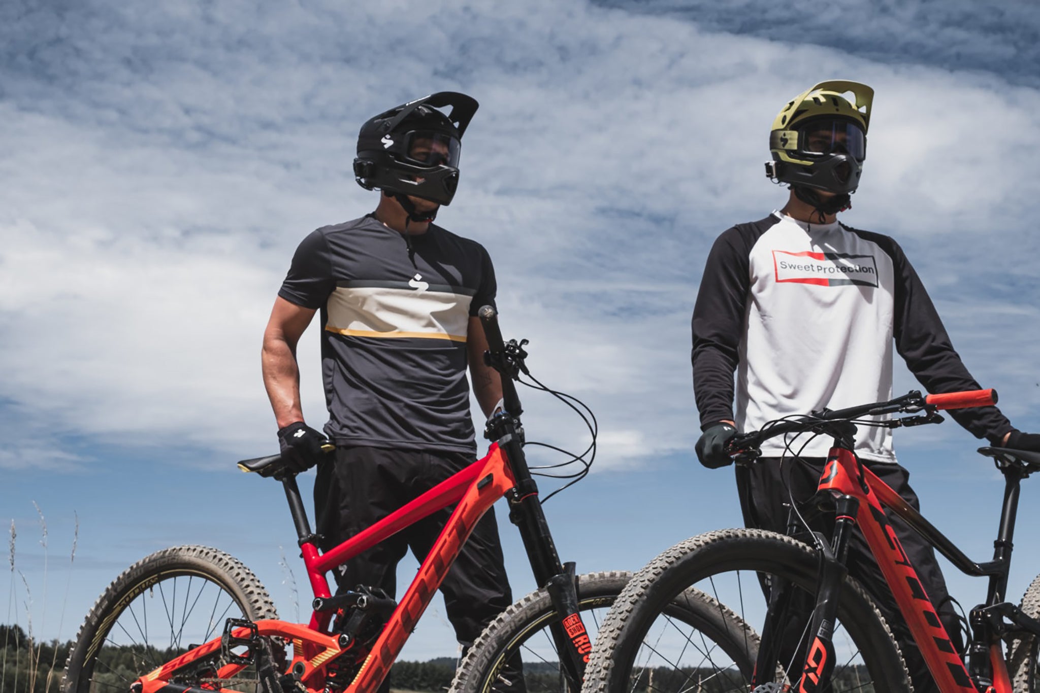Two mountain bikers wearing helmets and standing next to each other while holding their mountain bikes, using the Cardo Packtalk OUTDOOR Communication System to talk with each other