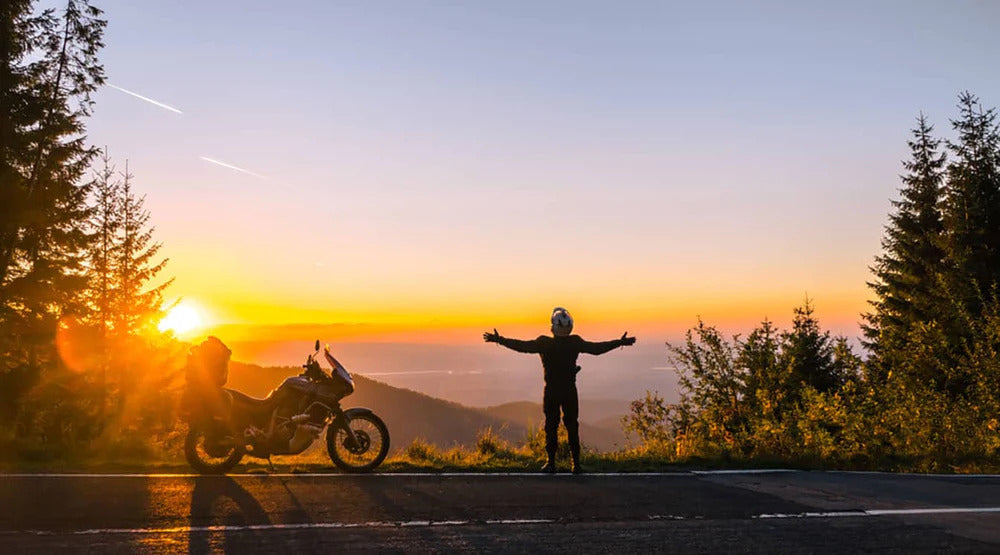 A motorcyclist in silhouette is standing beside his motorbike, arms spread, looking at a purple and orange sunset